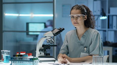 Portrait Of Young Beautiful Female Scientist In Uniform And Glasses Sitting At Desk In