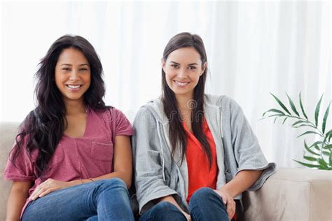 Two Smiling Women Sit On The Couch Together While Looking At The Stock Photo Image Of