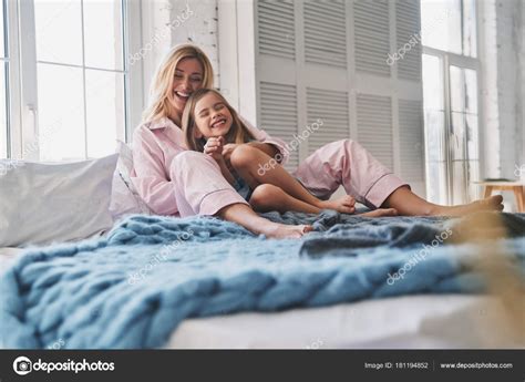 Скачать Красивая молодая мать обнимает дочь и улыбается сидя на кровати дома — стоковое