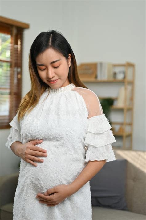 portrait beautiful asian pregnant woman stands in living room touching her belly with love