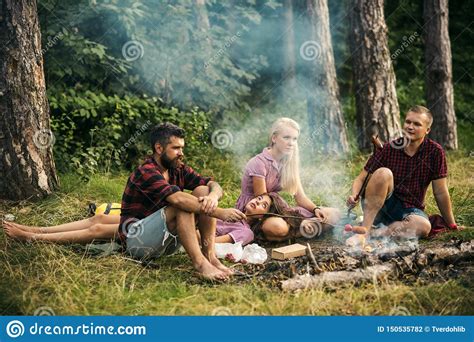 Two Couples Camping In Woods Men Cooking Sausages On Campfire Tired