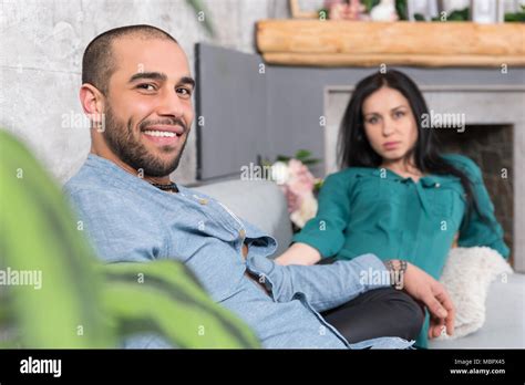 smiling international couple of man with beard and his brunette wife sitting on the sofa in the