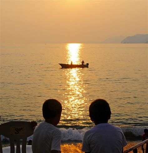 Two Boys Watching The Sun Set As A Fishing Boat Passes Through