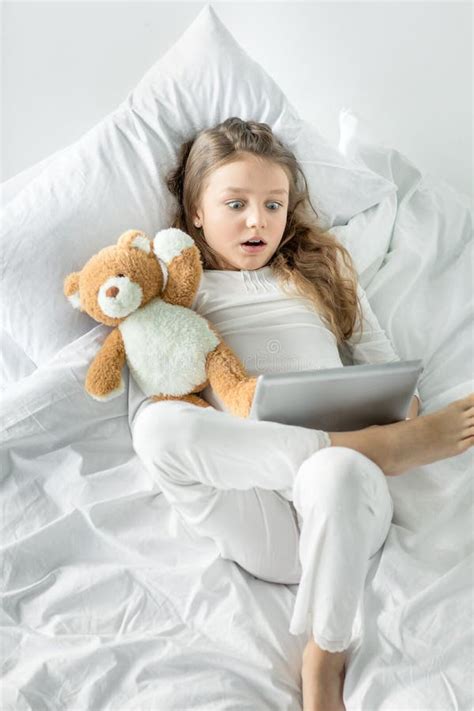 Little Girl In Pajamas Using Digital Tablet While Lying In Bed Stock