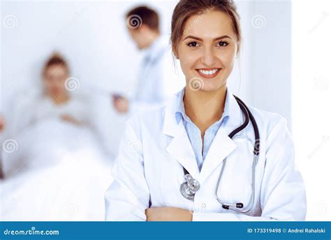 Female Doctor Smiling On The Background With Patient In The Bed And Two