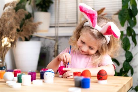 A Little Blonde Girl Sits At The Table And Paints Easter Eggs With