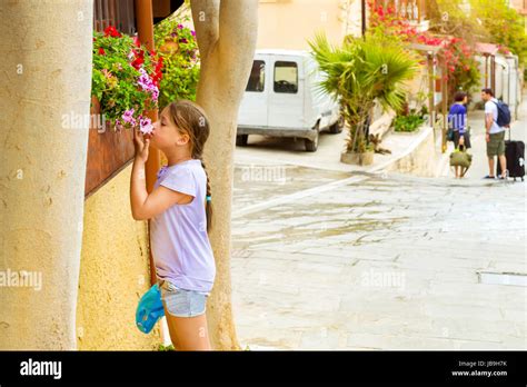 Cute Teenage Girl Sniffing Flowers Growing In A Flower Bed On Narrow Cobbled Street Tourists