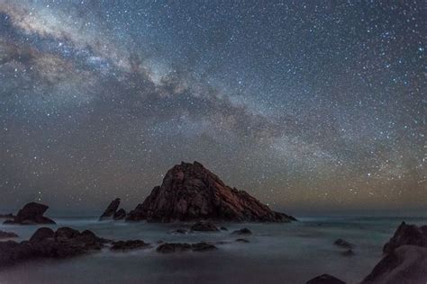 The Night Sky Is Filled With Stars And Clouds Above Rocks On An Ocean