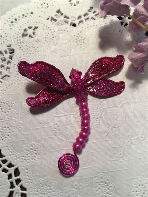 A Pink Dragonfly Brooch Sitting On Top Of A White Doily Next To Purple