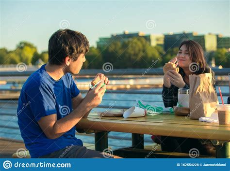 Two Young Adults Eating Hamburgers Outdoors By Lake Urban Setting