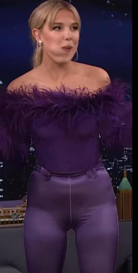 a woman wearing purple pants and an off the shoulder top is standing in front of a tv screen