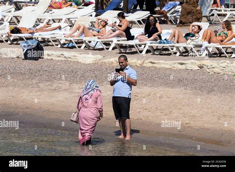 Muslim Woman Covered With Hijab On The Sea While Other Women Sunbathe Using Bikini In Eilat