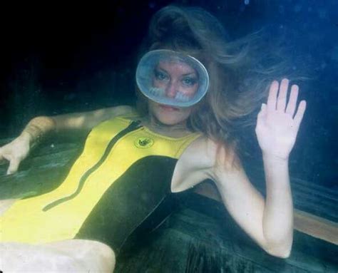 A Woman In A Yellow Wetsuit Floating Under Water With Her Hand Up To Her Face
