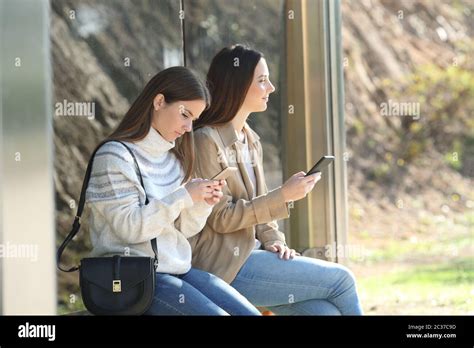 Two Women Waiting In A Bus Stop Checking Mobile Phones And Looking Away