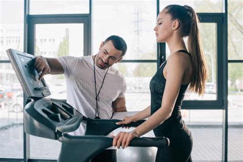 Male Personal Trainer Pointing At Treadmill Screen And Sportswoman