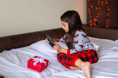 Little Brunette Girl In Pajamas Lies On The Bed At Home Looks Into The Digital Tablet Stock