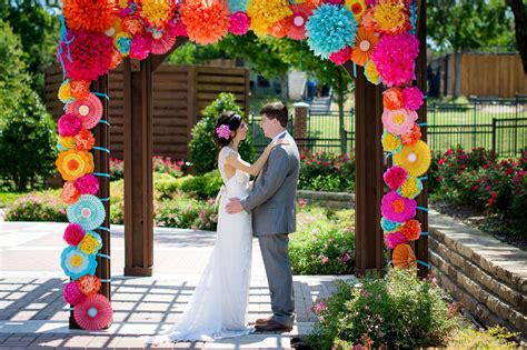 A Bride And Groom Standing Under An Arch Decorated With Paper Flowers