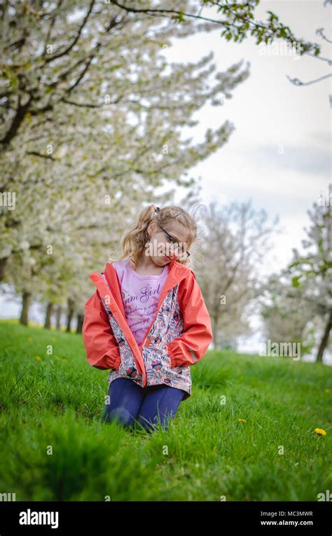 Adorable Blond Girl Sitting In Green Grass Under Trees With Blossoming