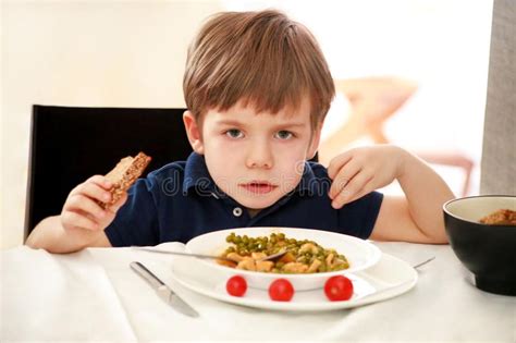Hungry Child Sitting In Chair At Table In Kitchen And Eating With Spoon