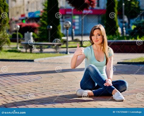 A Young And Pretty Girl In Jeans Sits On A Sidewalk Tile In The Middle