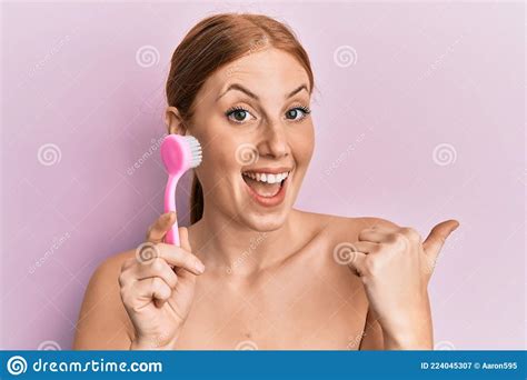 Young Irish Woman Using Facial Exfoliating Brush Pointing Thumb Up To The Side Smiling Happy