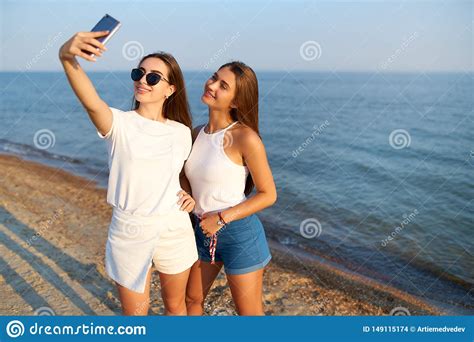 Two Young Women Taking A Selfie On The Beach With A Sea View Friends Are Smiling Looking At The