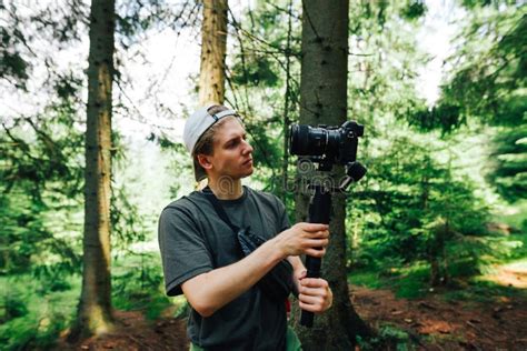 A Young Man With A Camera On A Stabilizer Stands In The Woods And
