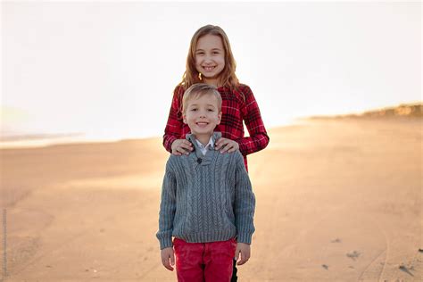 Big Sister Standing Behind Her Younger Brother On A Beach By Stocksy