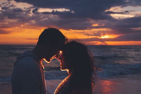 A Man And Woman Standing Next To Each Other Near The Ocean At Sunset Or Sunrise