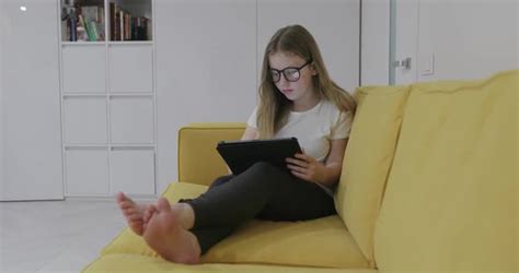 Barefoot Girl In Glasses Sitting On Sofa And Watching Video On Tablet While Resting At Home