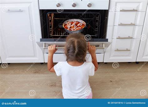 Charming Girl Sitting Near Gas Stove On Floor In Kitchen And Opening