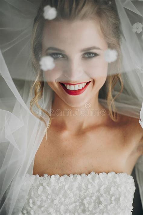 Stylish Happy Bride Posing Under Veil And Smiling In Soft Light Near