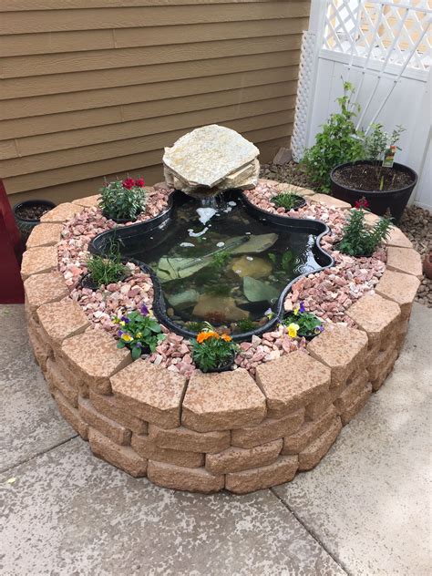 A Fish Pond In The Middle Of A Brick Wall With Flowers And Rocks Around It