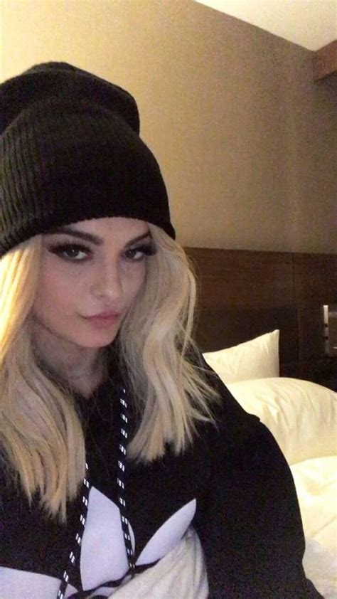 a woman with blonde hair wearing a beanie and black sweater sitting on a bed