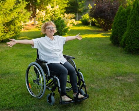 Happy Old Woman Spread Her Arms To The Sides While Sitting In A Wheelchair On A Walk Outdoors
