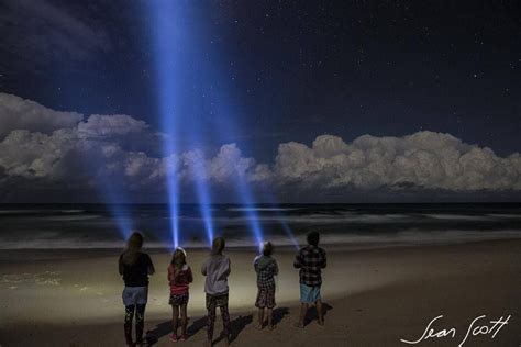 Four People Standing On The Beach Looking At An Object In The Sky With