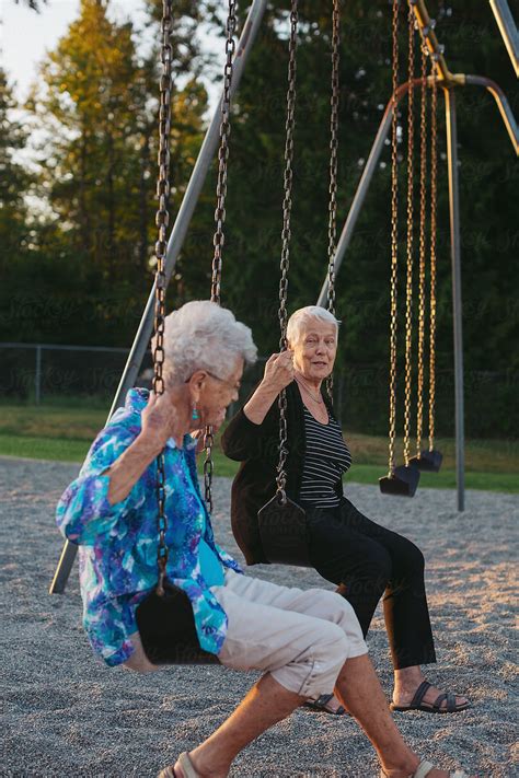 Two Happy Senior Caucasian Friends Swinging Together On Swingset At Sunset Del Colaborador De