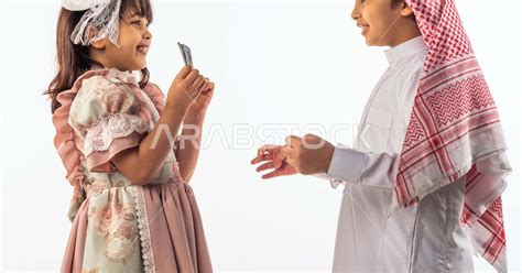 خلفية بيضاء لصبي وفتاه صغيران سعوديان مبتسمان ، تمسك الفتاه الصغيرة العيدية بيدها ينظر الطفل