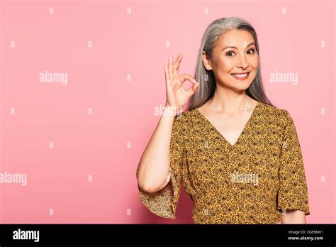 happy mature woman in yellow blouse with pattern showing okay gesture on pink background stock