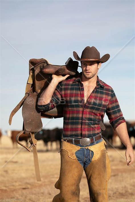A Man In Plaid Shirt And Cowboy Hat Holding Up A Saddle