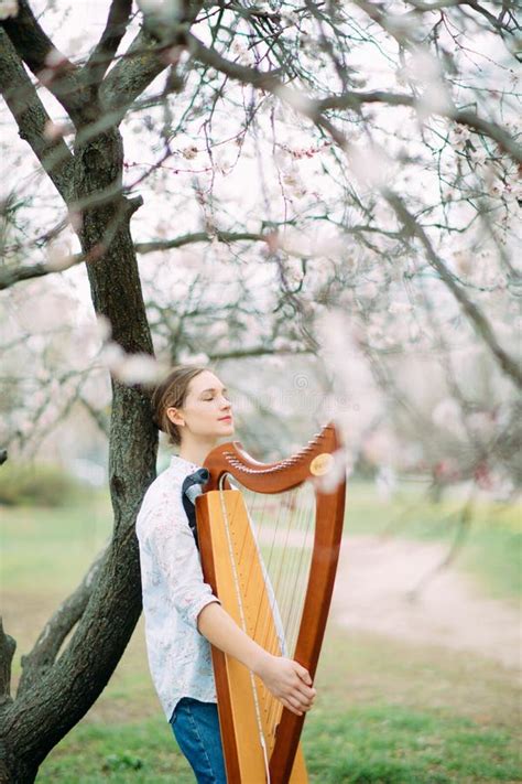 Woman Harpist Walks At Flowering Garden And Plays Harp Stock Photo