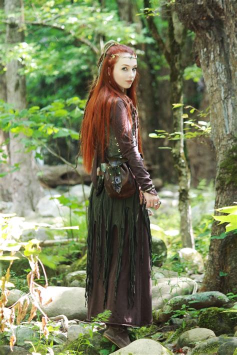 A Woman With Long Red Hair Standing In The Woods Wearing A Brown Dress