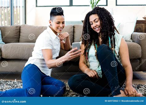 Smiling Lesbian Couple Sitting On Rug And Looking At Their Mobile Phone