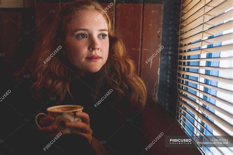 Thoughtful Redhead Woman Looking Through Window Blind In Cafe Casual