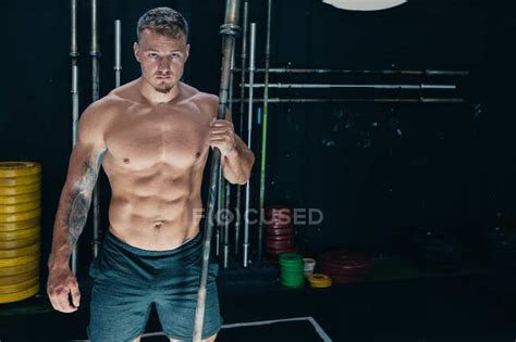 entschlossener mann mit athletischem nackten körper der mit metallhantel in dunkler turnhalle