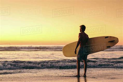 Man Standing On Beach At Sunrise Holding Surfboard San Diego