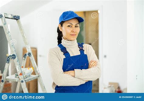 Portrait Of Positive Builder Woman In Blue Overalls Next To Stepladder