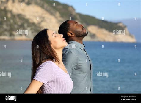 Side View Portrait Of A Happy Interracial Couple Breathing Fresh Air On The Beach Stock Photo