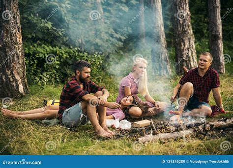 Two Couples Camping In Woods Men Cooking Sausages On Campfire Tired Brunette Lying On Blond