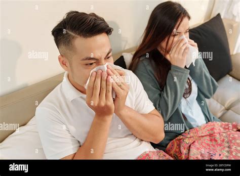 Young Sick Asian Couple Sitting In Bed Under Blanket And Blowing Noses